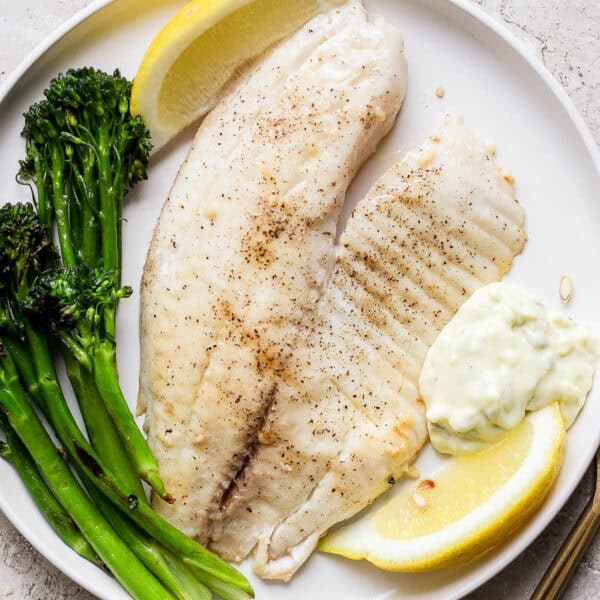 A plate with a piece of grilled tilapia, broccolini, lemon wedges and tartar sauce.