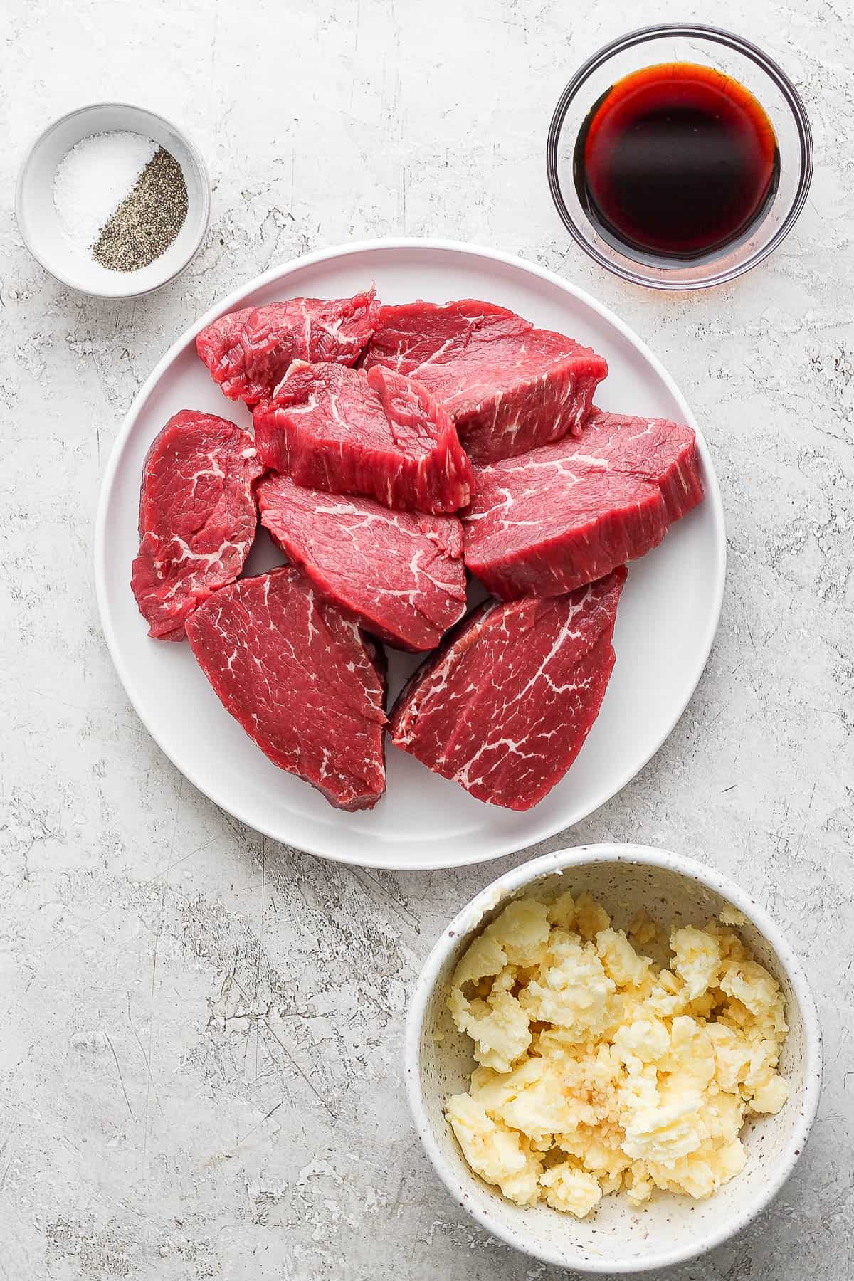 Raw steaks on a white plate next to seasoning, soy sauce, and garlic butter in separate bowls.