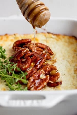 Side angle of a small casserole pan filled with baked goat cheese with pecans and fresh thyme on top with someone drizzling hot honey on top.