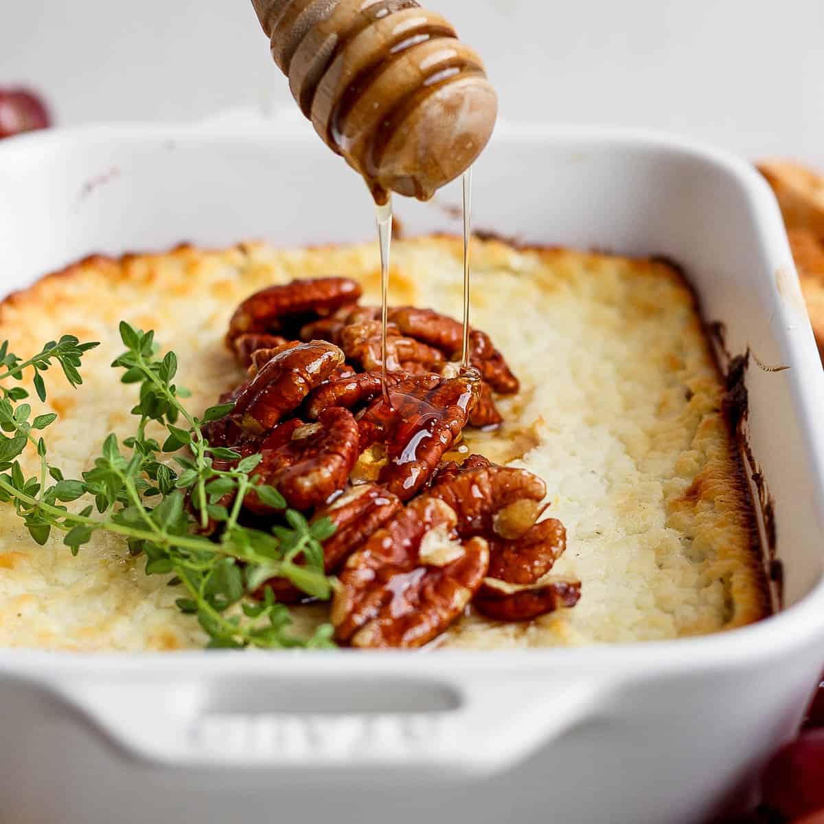 Side angle of a small casserole pan filled with baked goat cheese with pecans and fresh thyme on top with someone drizzling hot honey on top.