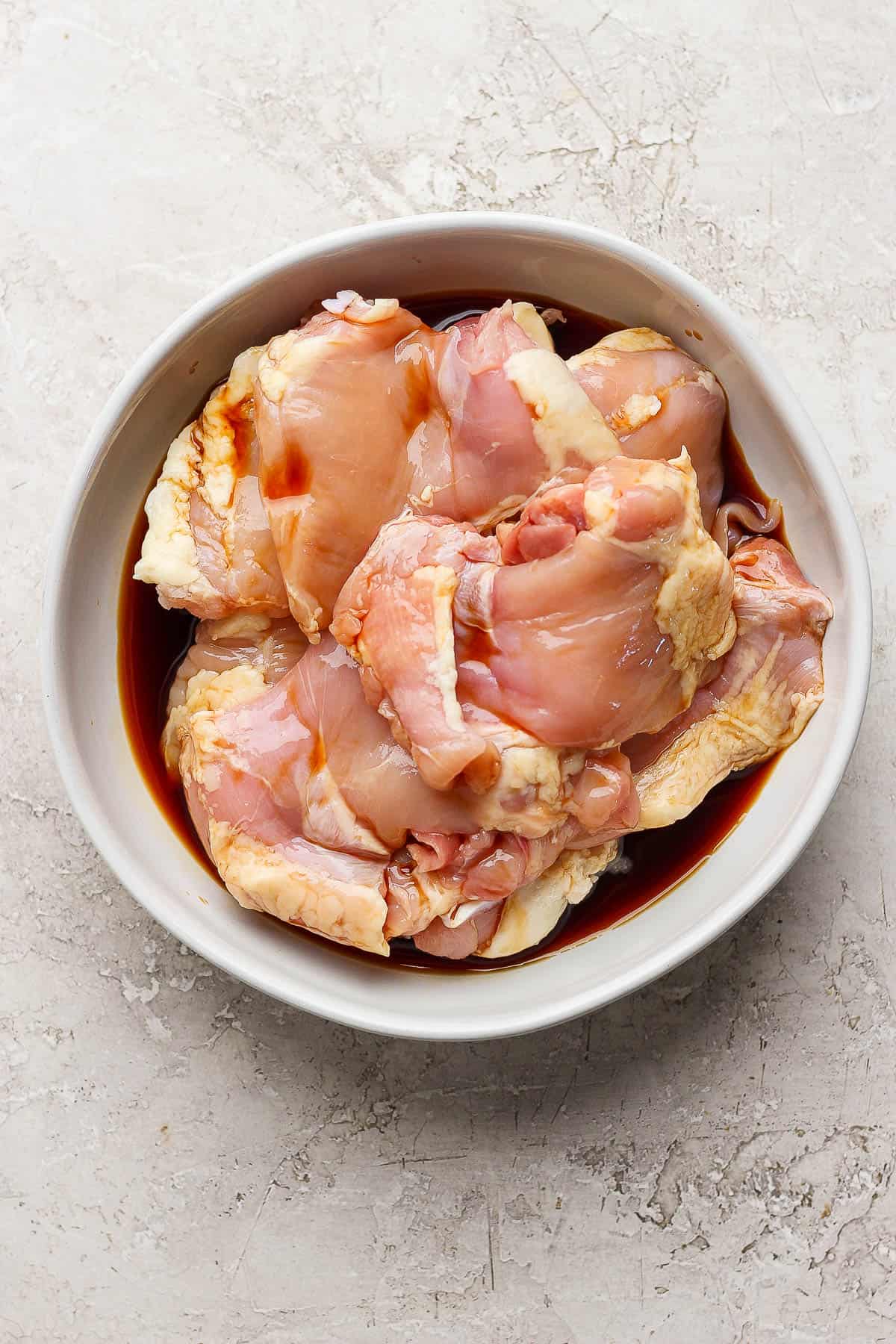 Raw chicken thighs marinating in a white bowl with soy sauce.