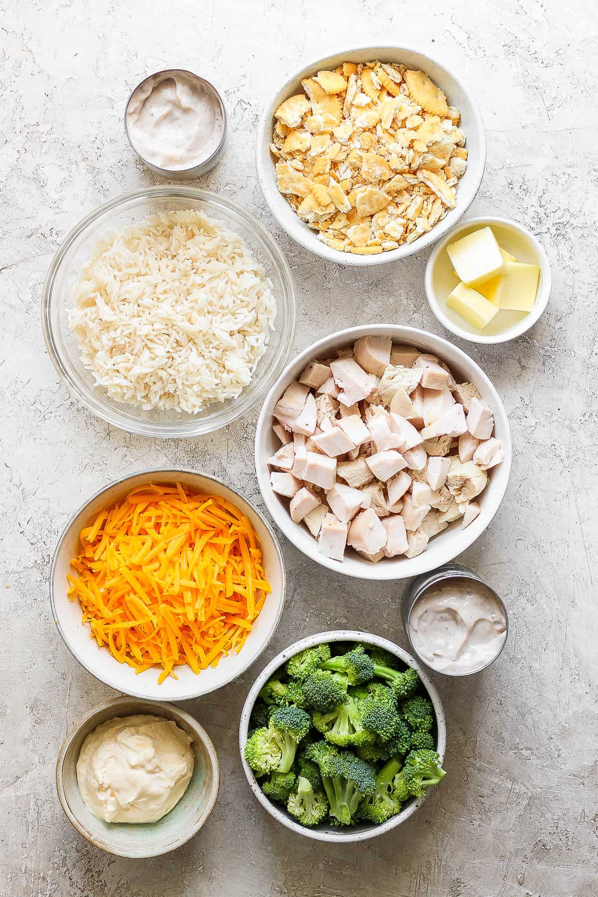 Individual bowls of crushed ritz crackers, cooked rice, butter, cubed chicken, shredded cheddar cheese, cream of mushroom soup, broccoli florets, and mayo.