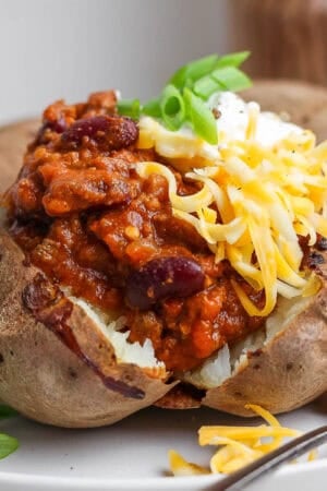 Straight on shot of a baked potato cut open on a plate and topped with chili, cream cheese, shredded cheese and green onion slices.