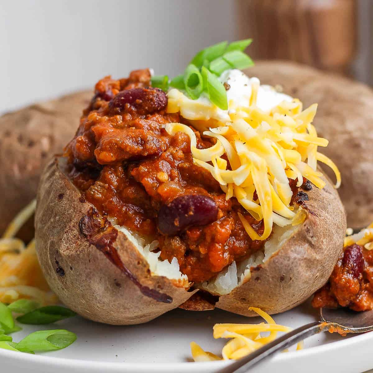 Straight on shot of a baked potato cut open on a plate and topped with chili, cream cheese, shredded cheese and green onion slices.