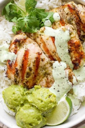 A bowl of rice with grilled cilantro lime chicken with cilantro lime sauce on top, guacamole, lime wedges and fresh cilantro.