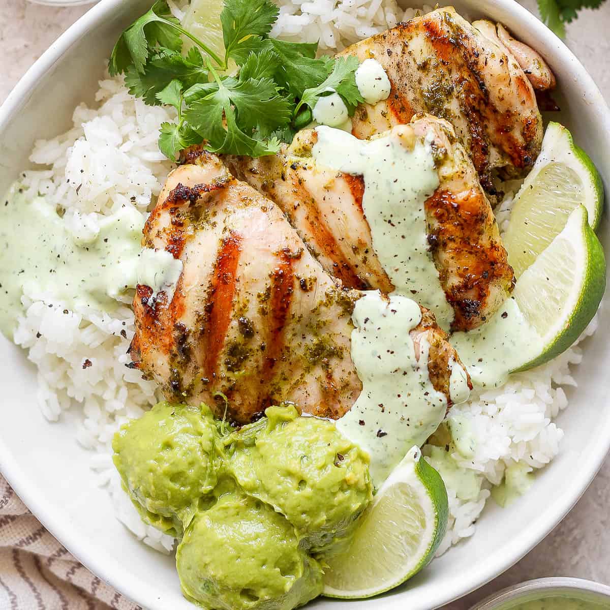 A bowl of rice with grilled cilantro lime chicken with cilantro lime sauce on top, guacamole, lime wedges and fresh cilantro.