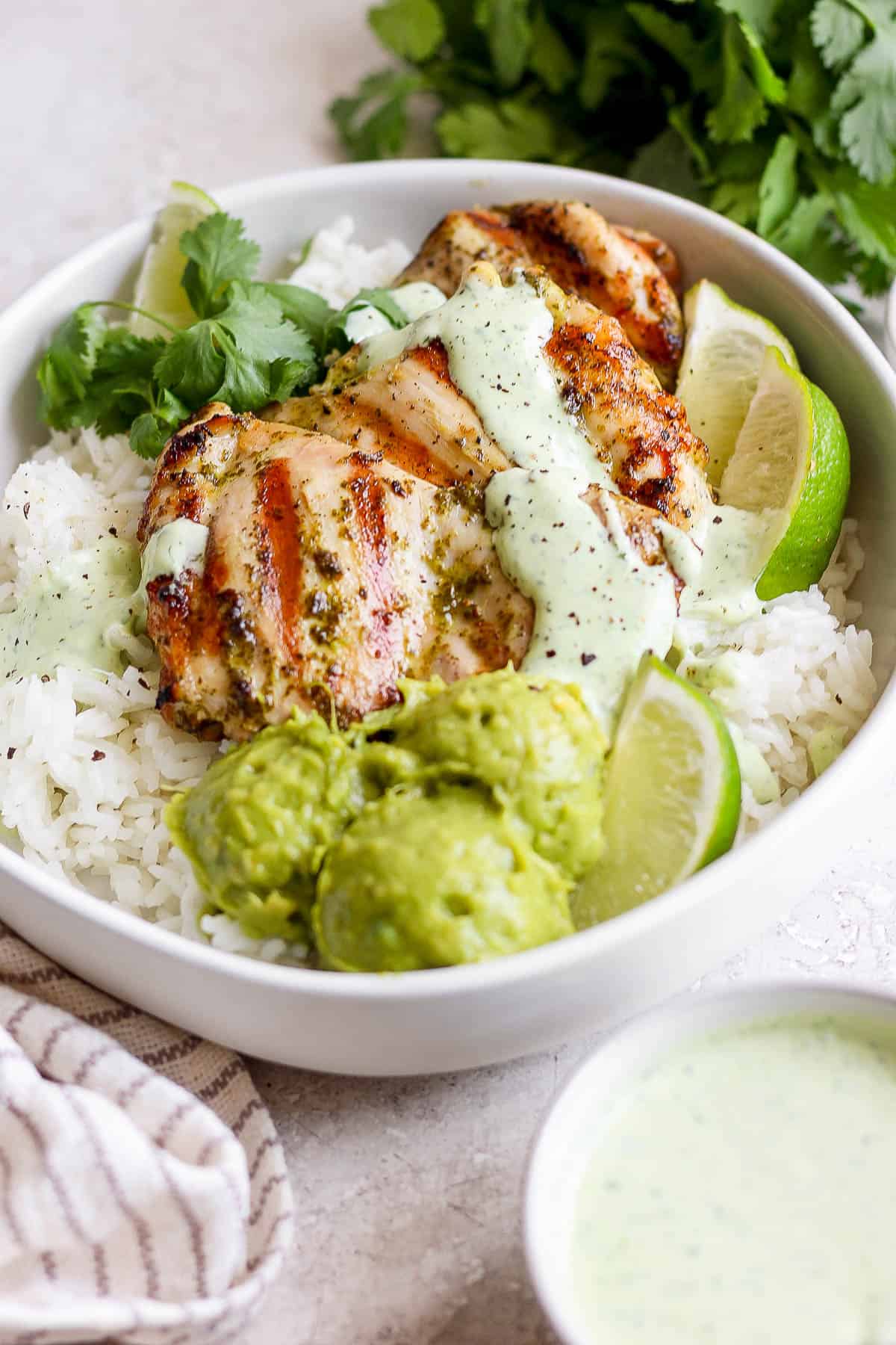 Cooked chicken on some white rice with guacamole, yogurt sauce, cilantro, and lime wedges.