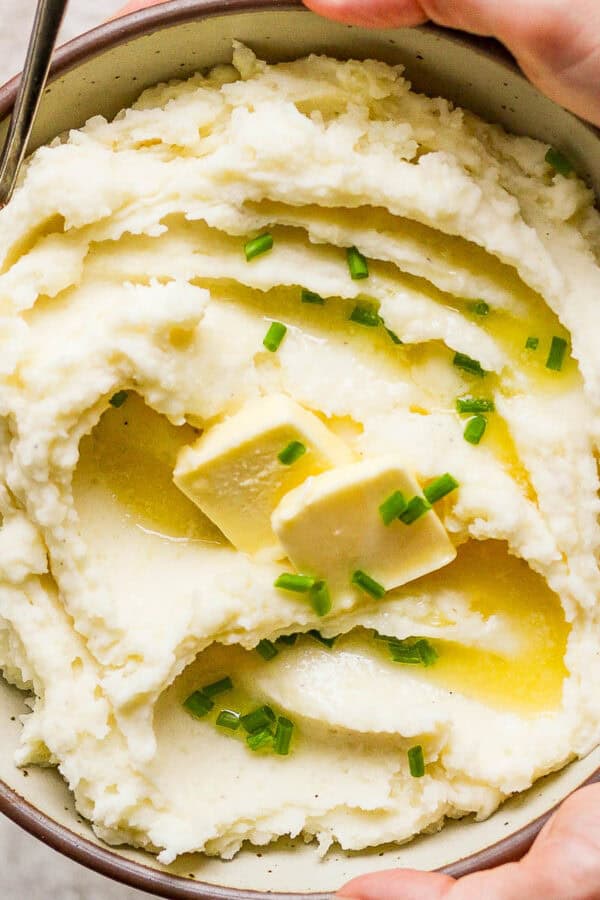 Someone holding a bowl of creamy mashed potatoes with extra butter and chives on top.