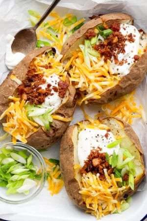 Top down shot of a platter of 3 loaded baked potatoes with sour cream, cheese, bacon and green onions on top.