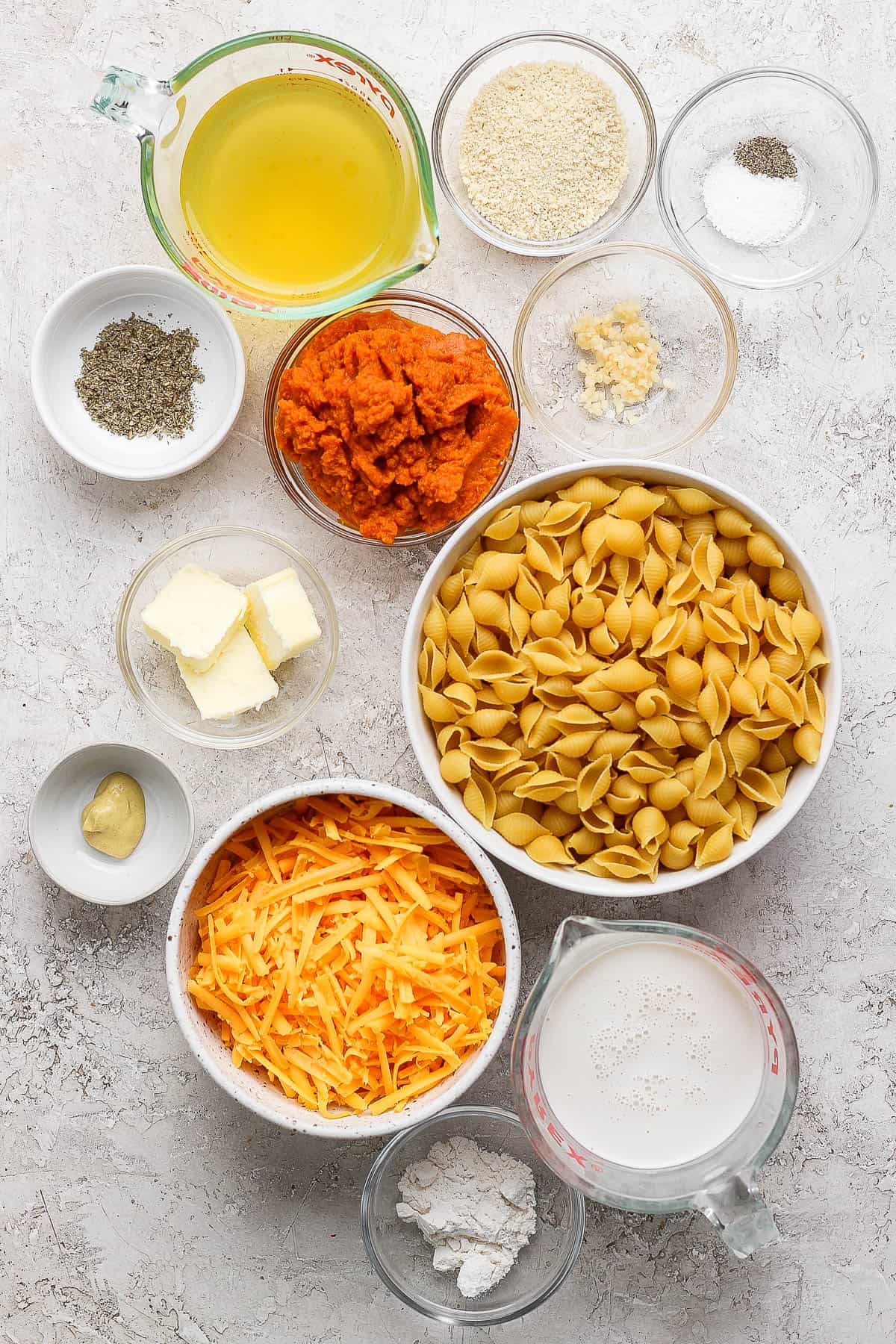 Individual bowls of flour, milk, shredded cheese, mustard, pasta shells, butter, pumpkin, seasonings, minced garlic, salt, pepper, bread crumbs, and chicken stock.