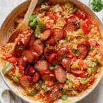 Top down shot of a braiser pan filled with sausage, rice, bell peppers and tomatoes with a wooden spoon sticking out.