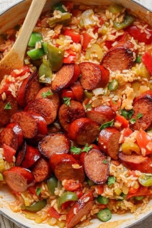 Top down shot of a braiser pan filled with sausage, rice, bell peppers and tomatoes with a wooden spoon sticking out.