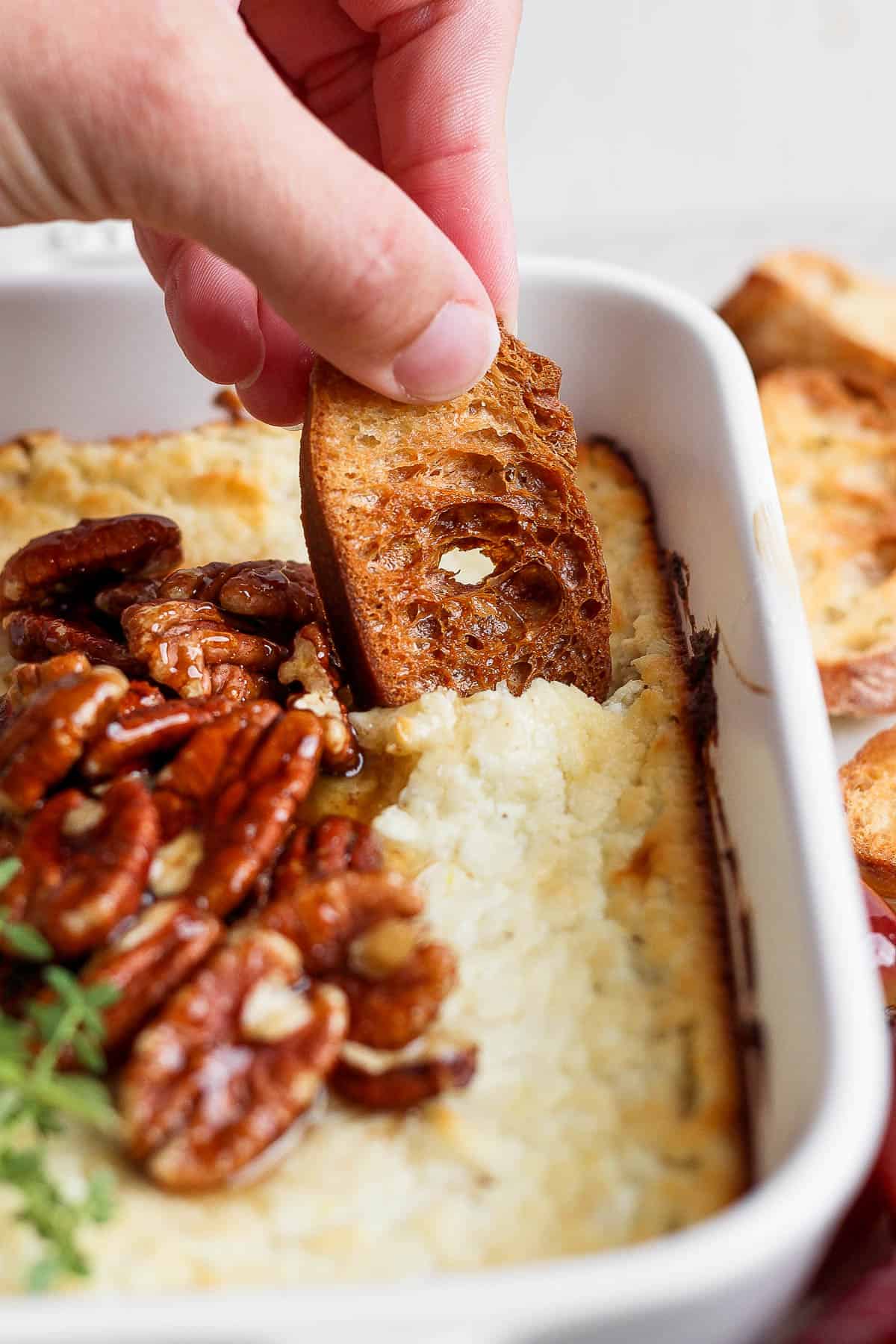 A hand scooping up baked goat cheese with a crostini.