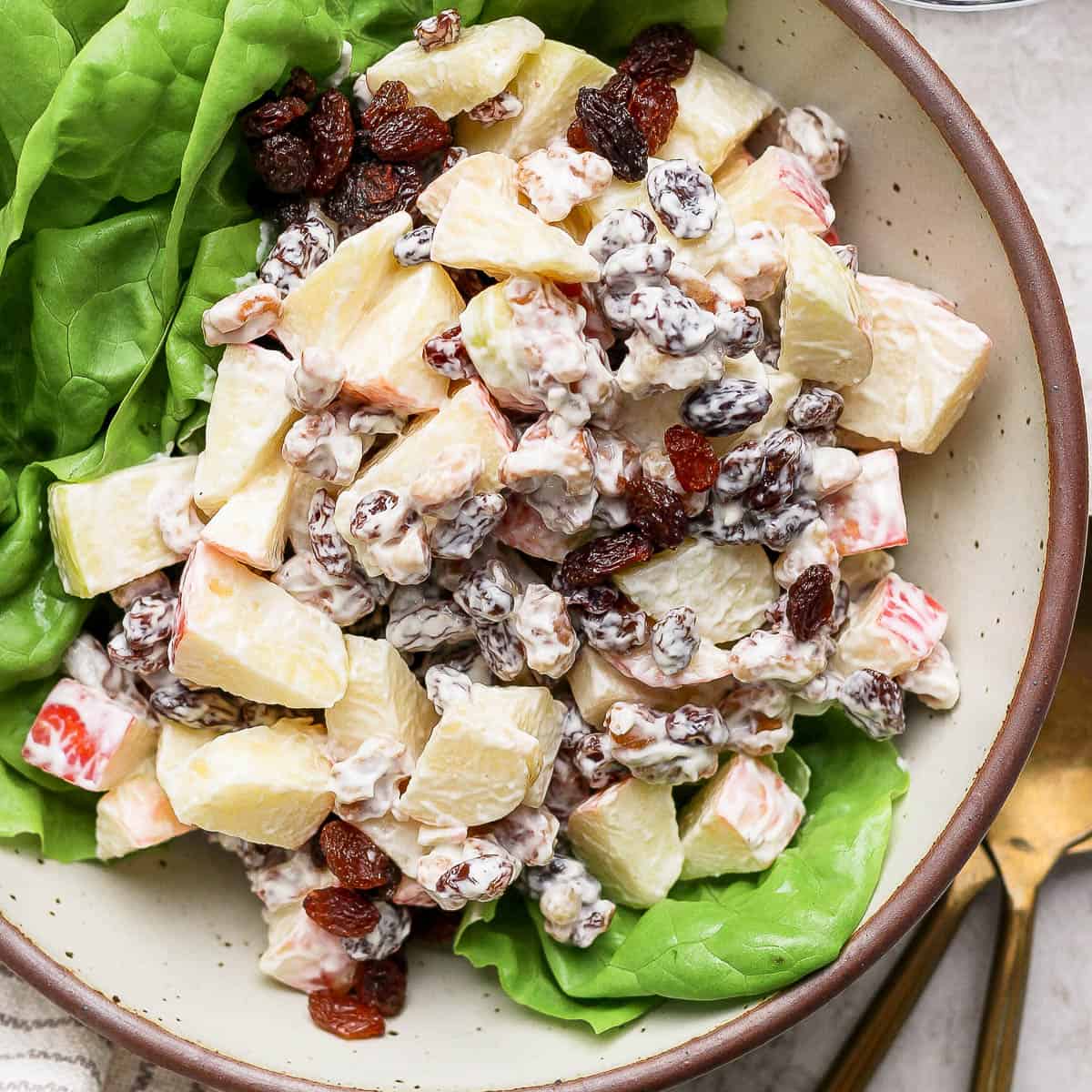 Top down shot of a bowl of homemade apple salad on top of butter lettuce with a gold spoon and fork next to the bowl.