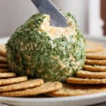 A cheese ball rolled in dried parsley on a plate with crackers and a small butter knife scooping some out.