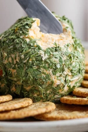 A cheese ball rolled in dried parsley on a plate with crackers and a small butter knife scooping some out.