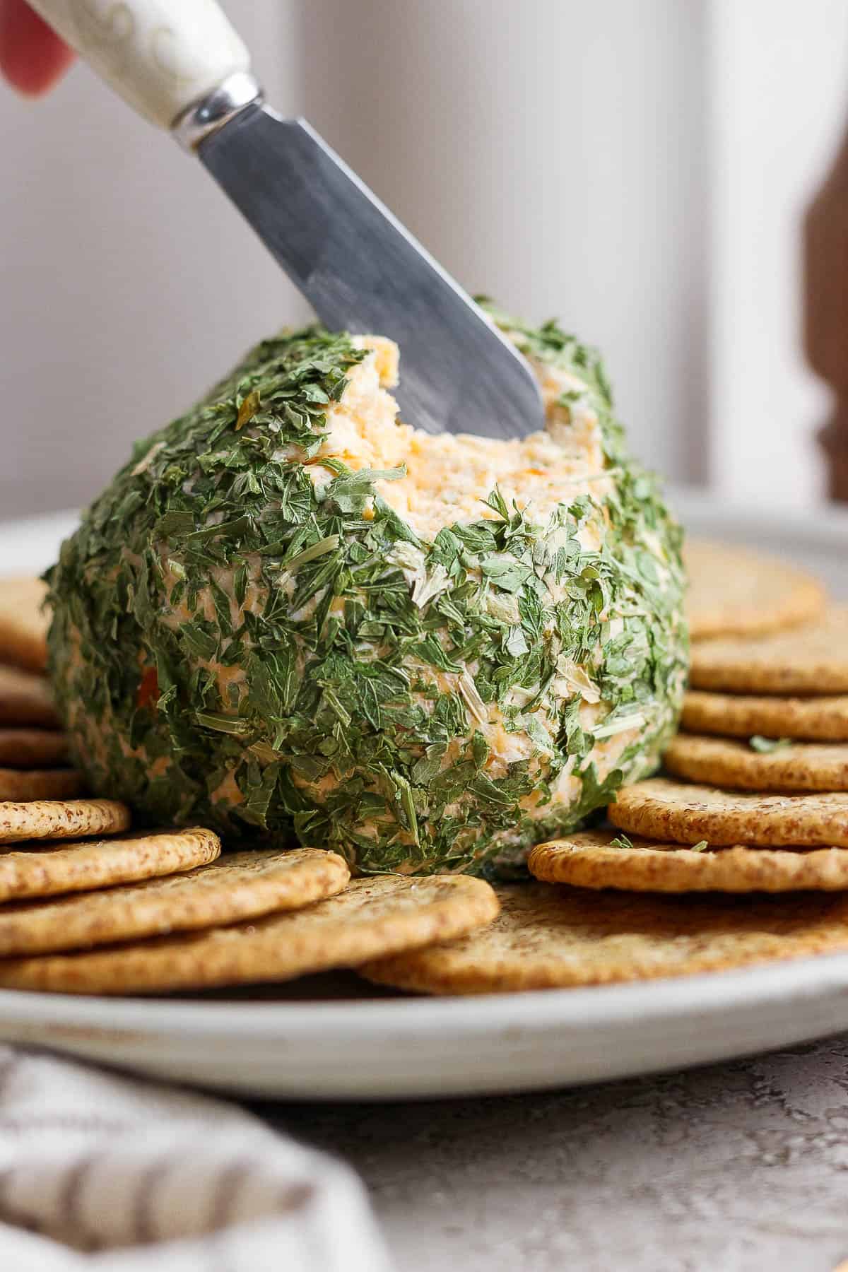 A cheese ball on a plate surrounded by crackers with a knife scooping out some cheese from the ball.