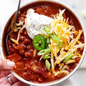 Someone holding a bowl of smoked chili with sour cream, shredded cheese, green onion slices and a slice of jalapeno.