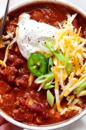 Someone holding a bowl of smoked chili with sour cream, shredded cheese, green onion slices and a slice of jalapeno.