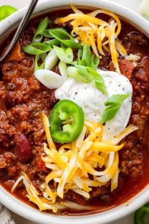 Top down shot of a bowl of bison chili with sour cream, green onion, shredded cheese and a slice of jalapeno with a spoon sticking out.