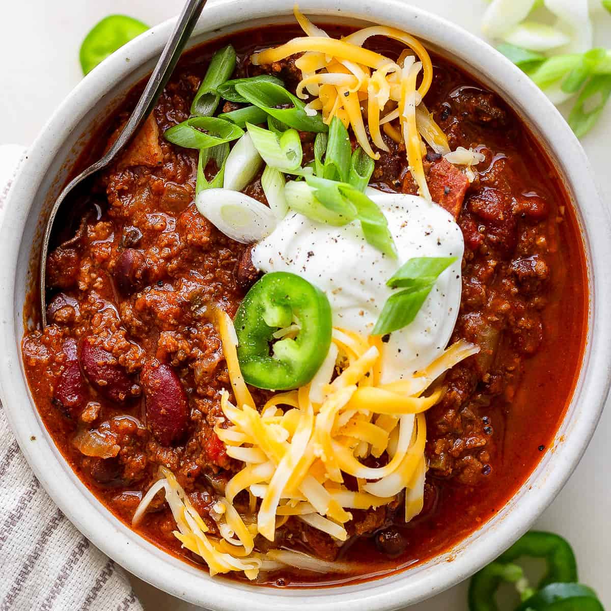 Top down shot of a bowl of bison chili with sour cream, green onion, shredded cheese and a slice of jalapeno with a spoon sticking out.