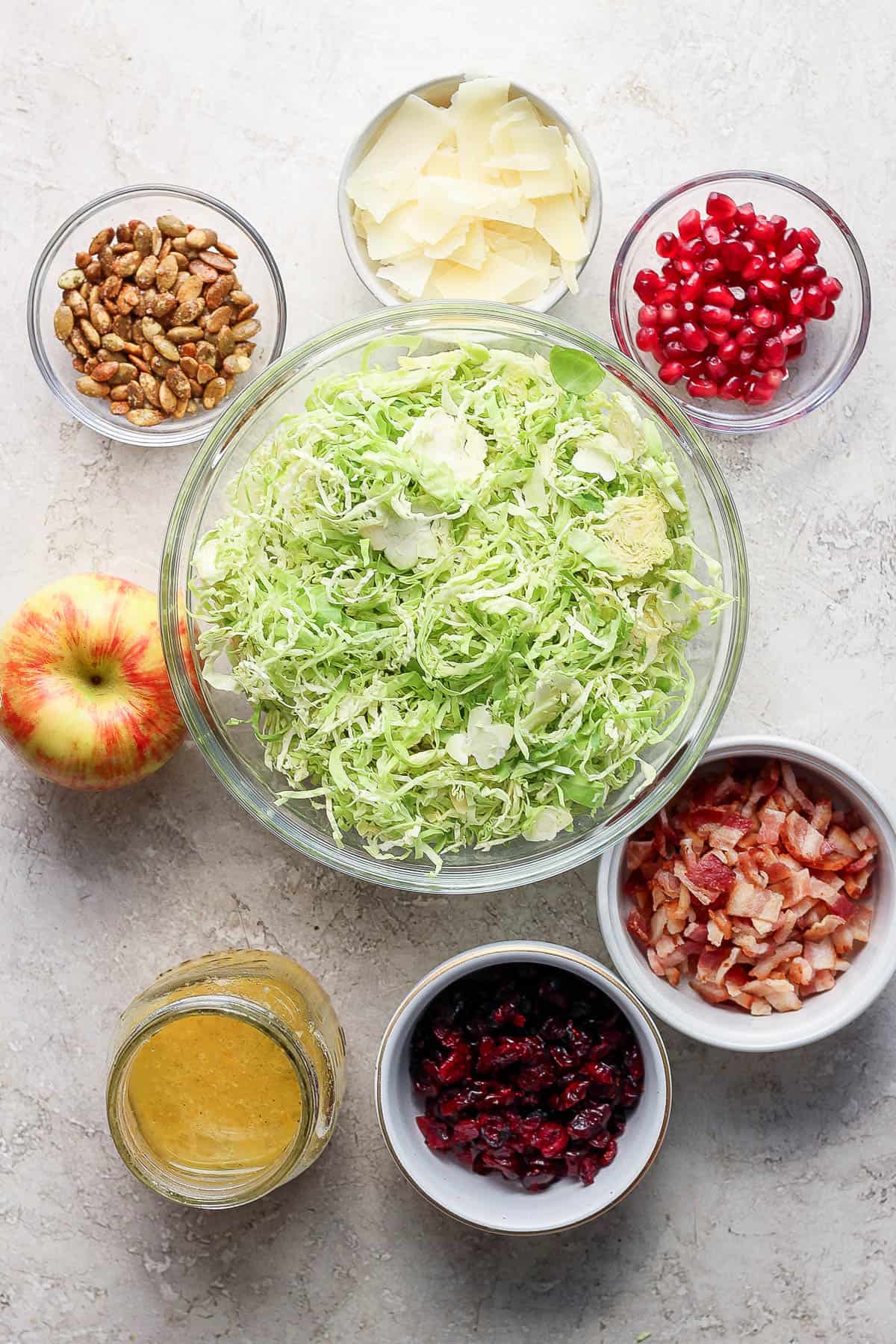 Individual bowls of shaved brussel sprouts, pomegranites, pepitas, chopped bacon, cranberries, a dressing, and a whole apple.