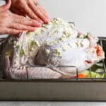 A raw turkey sitting in a roasting pan with someone rubbing herbed butter all over it.