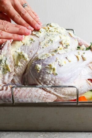 A raw turkey sitting in a roasting pan with someone rubbing herbed butter all over it.