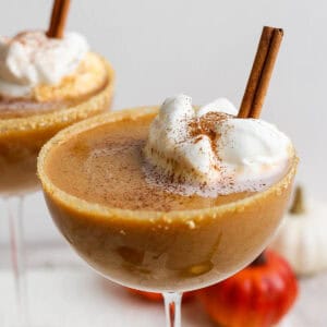 Close up shot of a coupe glass with a pumpkin pie martini in it with homemade whipped cream and a cinnamon stick in it with another one in the background.