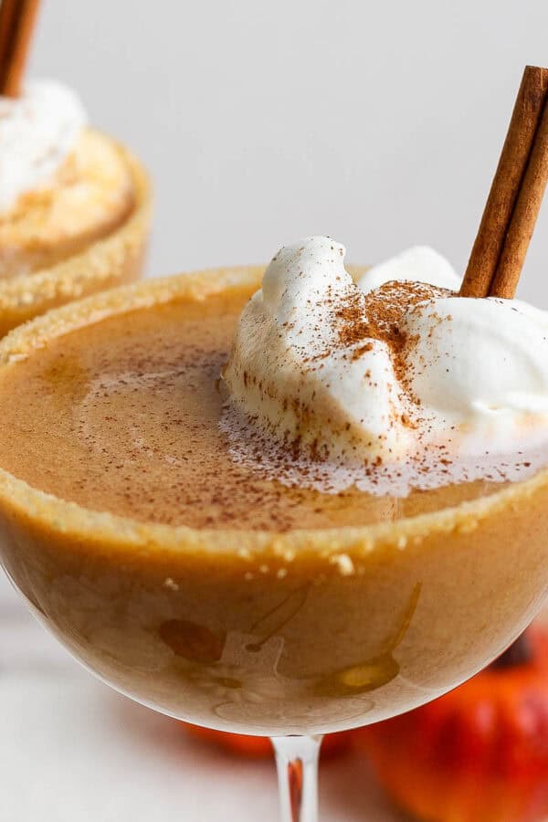 Two coupe glasses, each with a pumpkin pie martini in it, whipped cream and a cinnamon stick.