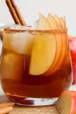 A glass filled with a homemade apple cider bourbon cocktail with ice, apple slices and a cinnamon stick in it.