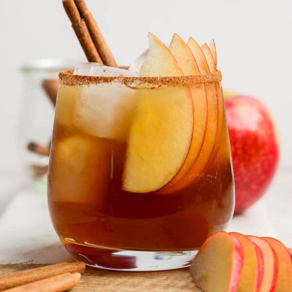 A glass filled with a homemade apple cider bourbon cocktail with ice, apple slices and a cinnamon stick in it.