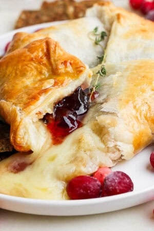 A plate filled with a baked brie in puff pastry with melted brie cheese coming out, cranberries around it with some crackers and fresh thyme.