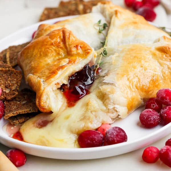 A plate filled with a baked brie in puff pastry with melted brie cheese coming out, cranberries around it with some crackers and fresh thyme.