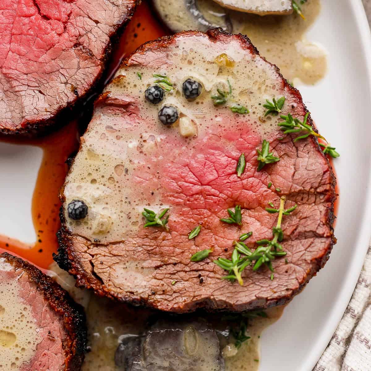 Top down shot of a slice of beef tenderloin with a creamy mushroom sauce over top, whole black peppercorns and fresh thyme.