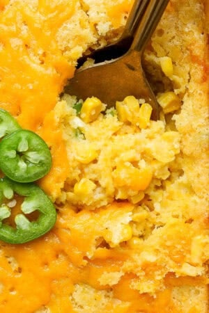 Top down, close up shot of a the corner of a pan of corn casserole with two spoons sticking out and three slices of jalapeno.