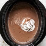 A crock pot fulled with homemade hot chocolate and whipped cream on top with shaved chocolate.