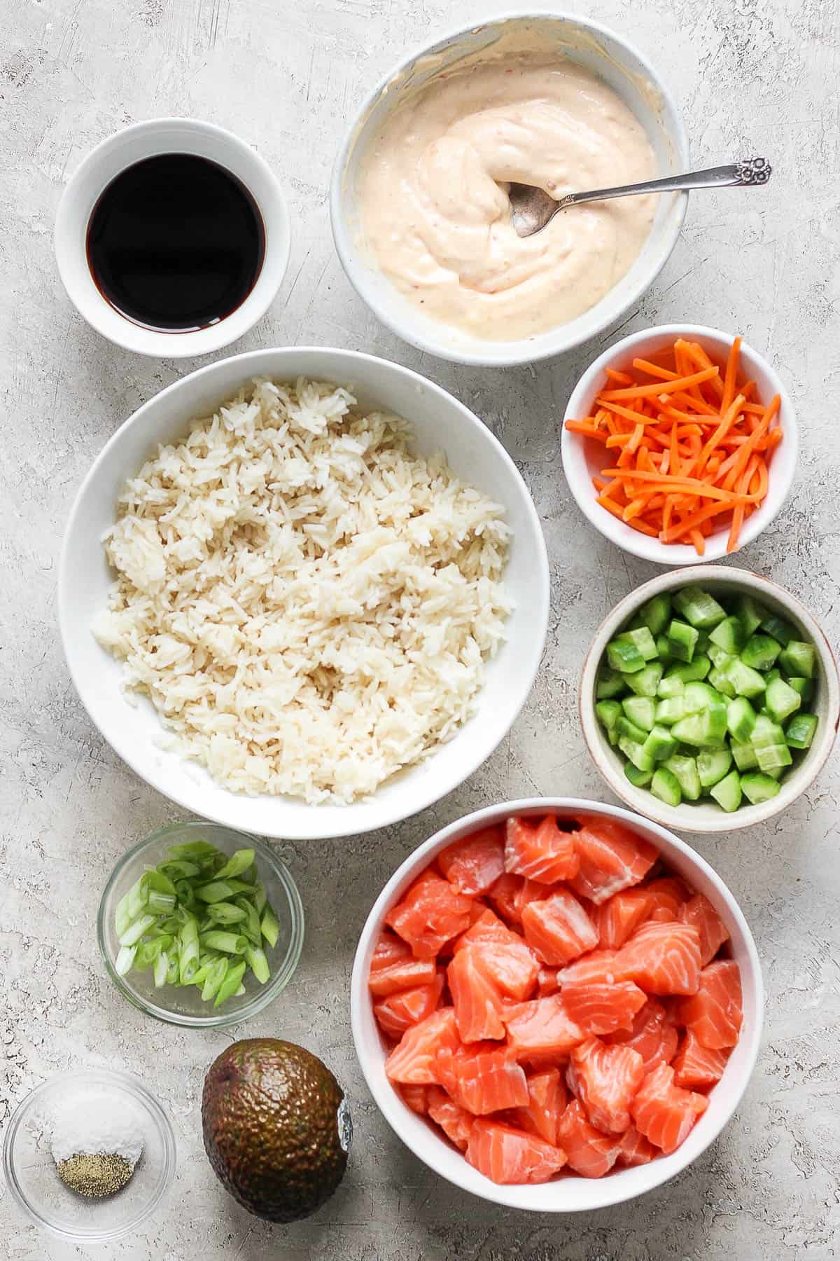 Individual bowls of soy sauce, bang bang sauce, cooked rice, carrot slices, sliced and quartered cucumber, raw salmon chunks, green onion, salt and pepper, and a whole avocado.