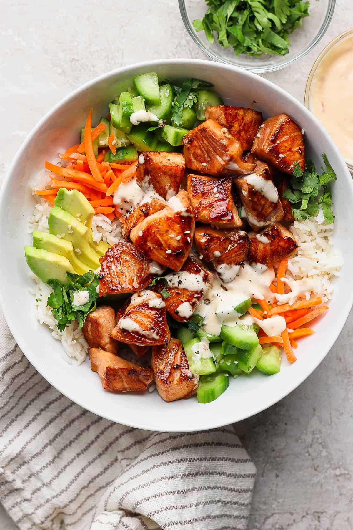 A bowl with rice, carrot sticks, cucumber, sliced avocado, salmon bites, bang bang sauce, and fresh cilantro.