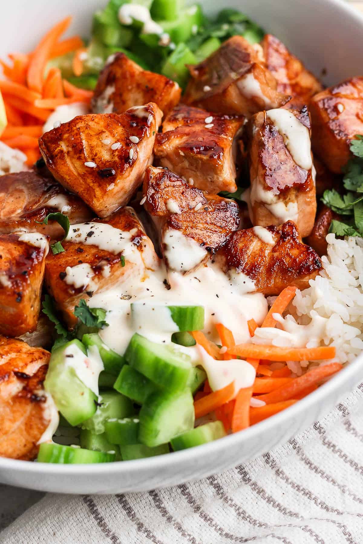 A bowl with rice, carrot sticks, cucumber, sliced avocado, salmon bites, bang bang sauce, and fresh cilantro.