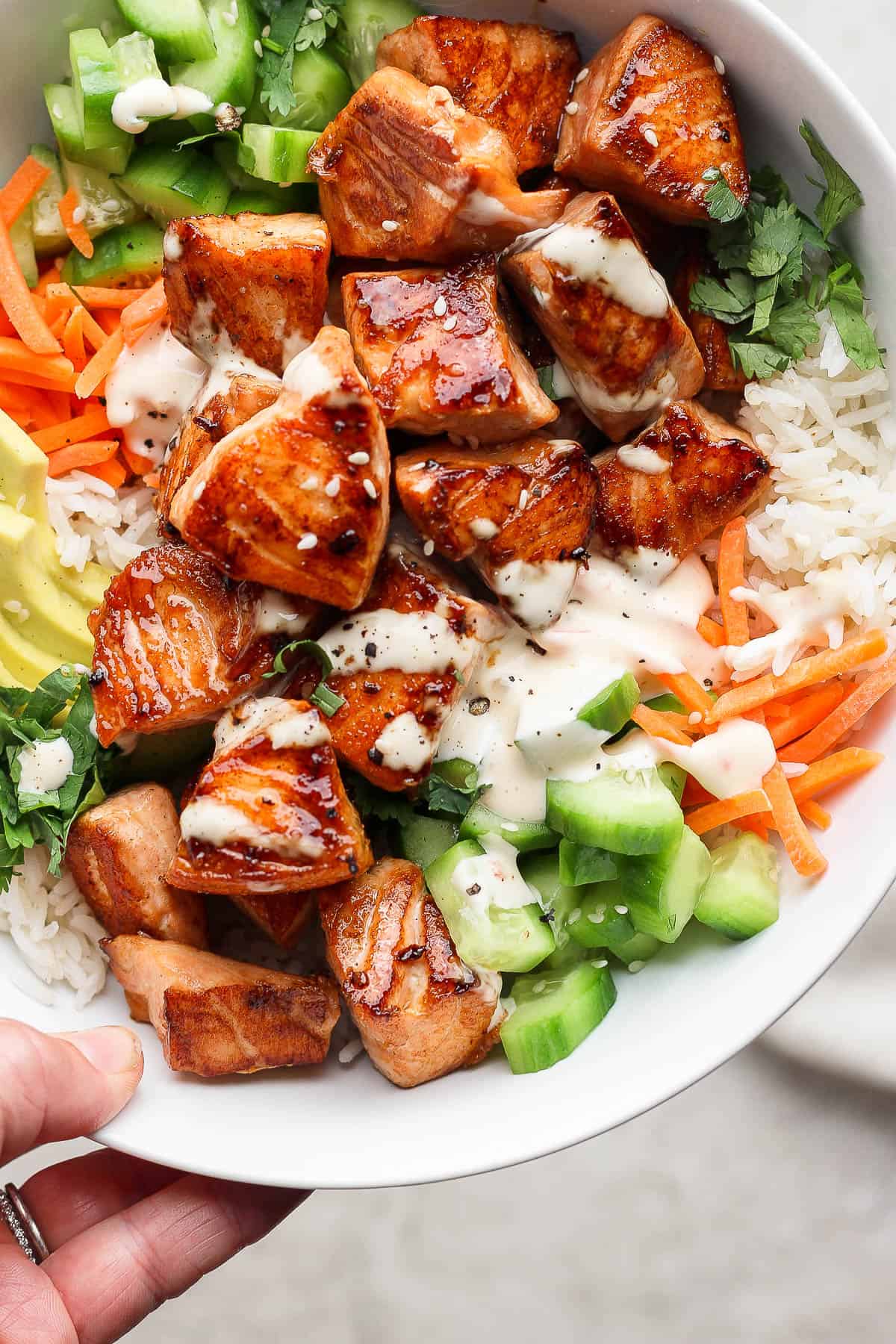 A bowl with rice, carrot sticks, cucumber, sliced avocado, salmon bites, bang bang sauce, and fresh cilantro.