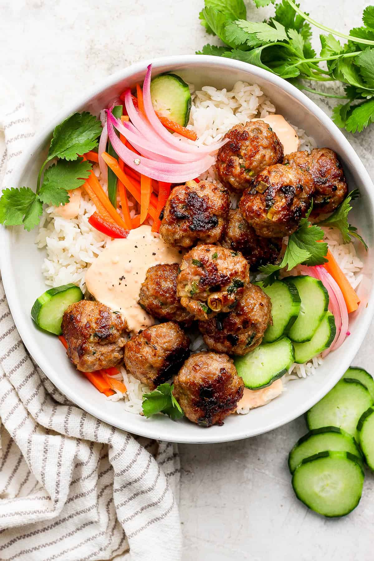 A bowl with rice, meabtalls, fresh cucumber, pickled veggies, cilantro, and spicy mayo.