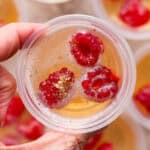 Someone holding a champagne jello shot with raspberries and gold sparkles inside.