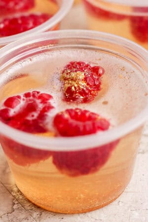 Side shot of a champagne jello shot with raspberries and gold sparkles inside.