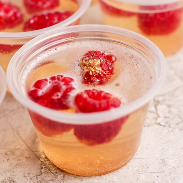 Side shot of a champagne jello shot with raspberries and gold sparkles inside.