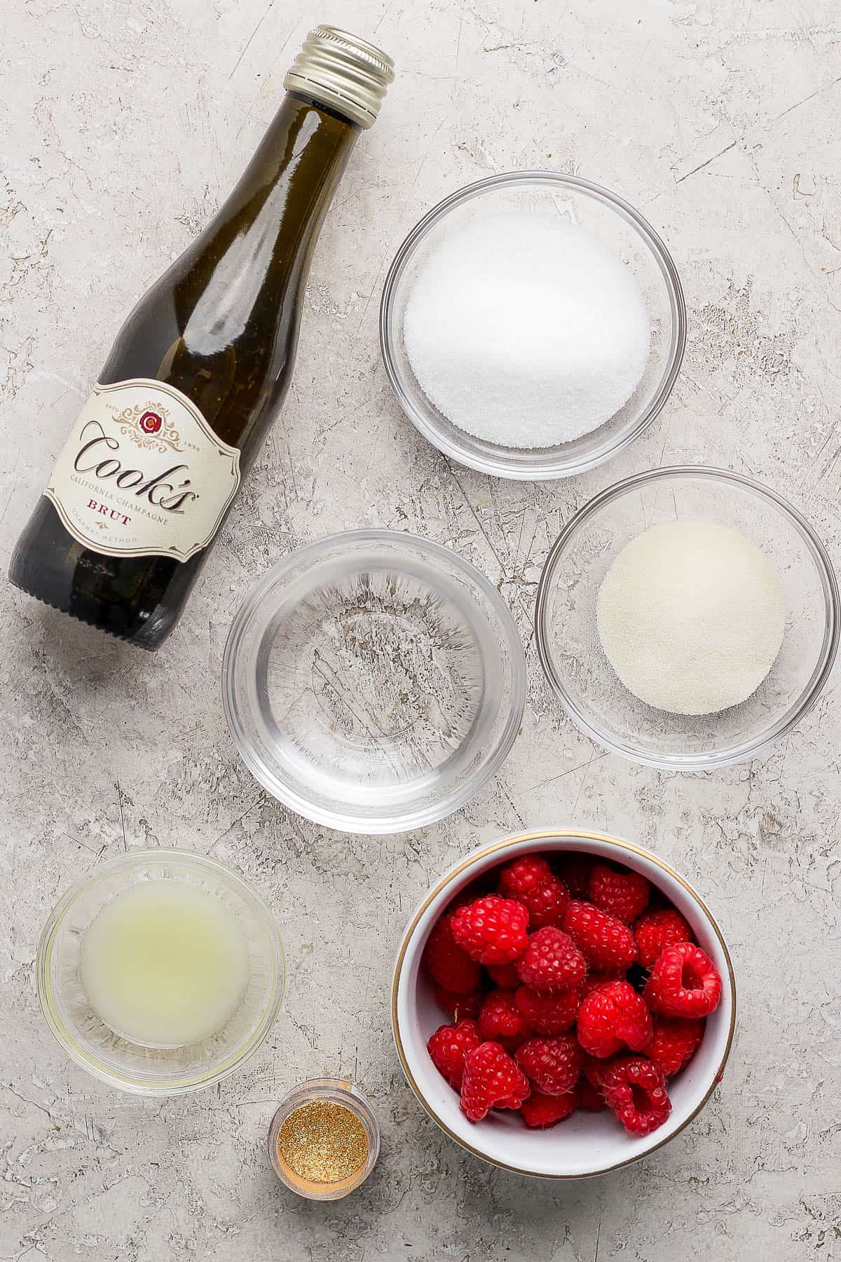 A bottle of Cook's champagne and individual bowls of sugar, gelatin, vodka, lemon juice, edible gold glitter, and raspberries.