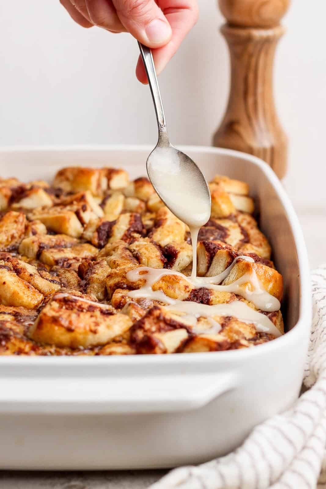 A hand holding a spoon drizzling icing over cinnamon roll casserole.