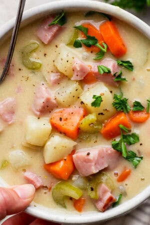 Someone holding a bowl of ham and potato soup with a spoon sticking out and parsley on top for garnish.