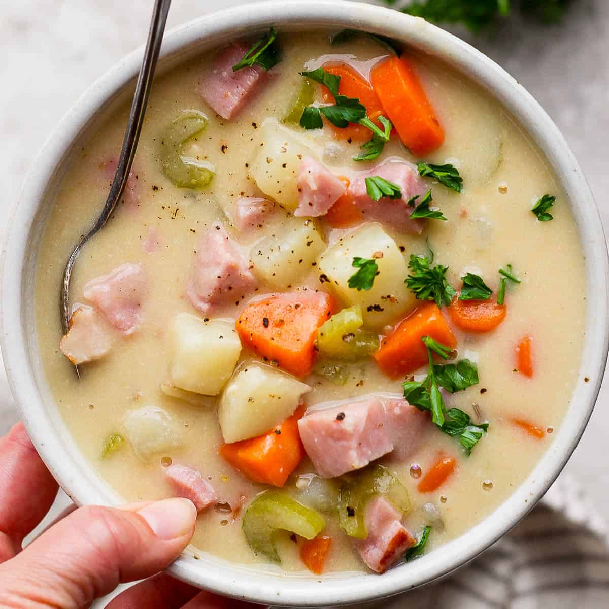 Someone holding a bowl of ham and potato soup with a spoon sticking out and parsley on top for garnish.