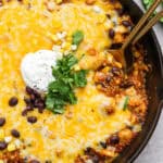 A cast iron skillet filled with a quinoa enchilada bake with sour cream and cilantro on top with two spoons sticking out.